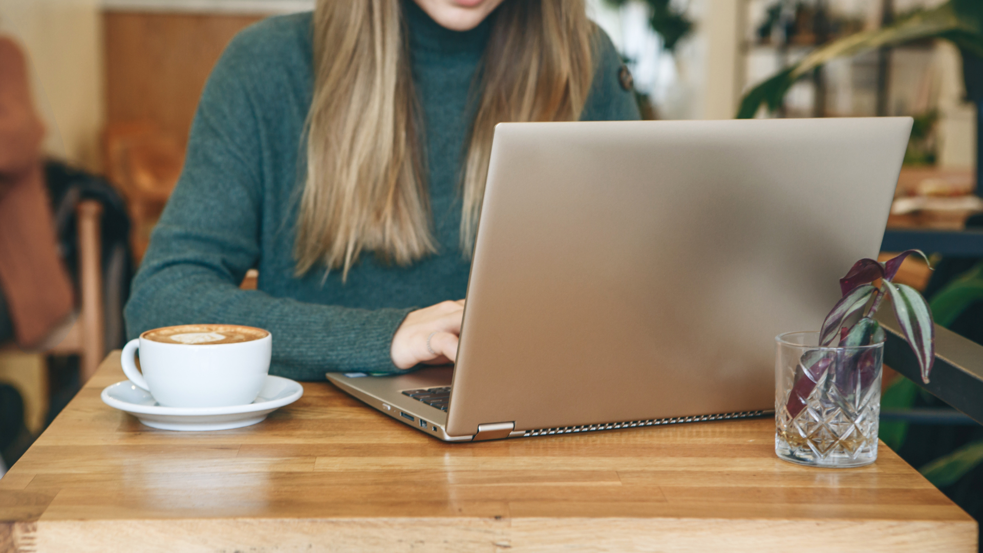 A person in a dark green turtleneck works on a laptop at a wooden table with a cup of coffee and a small glass vase.