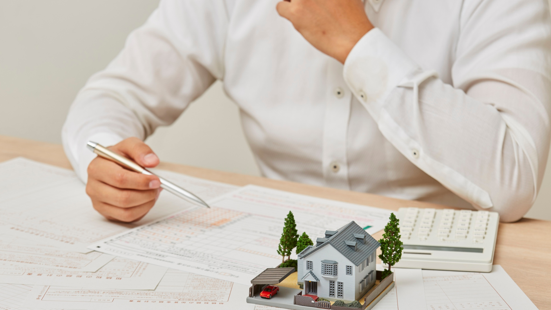 A person in a white shirt points with a pen at documents next to a miniature house model and a calculator on a desk.