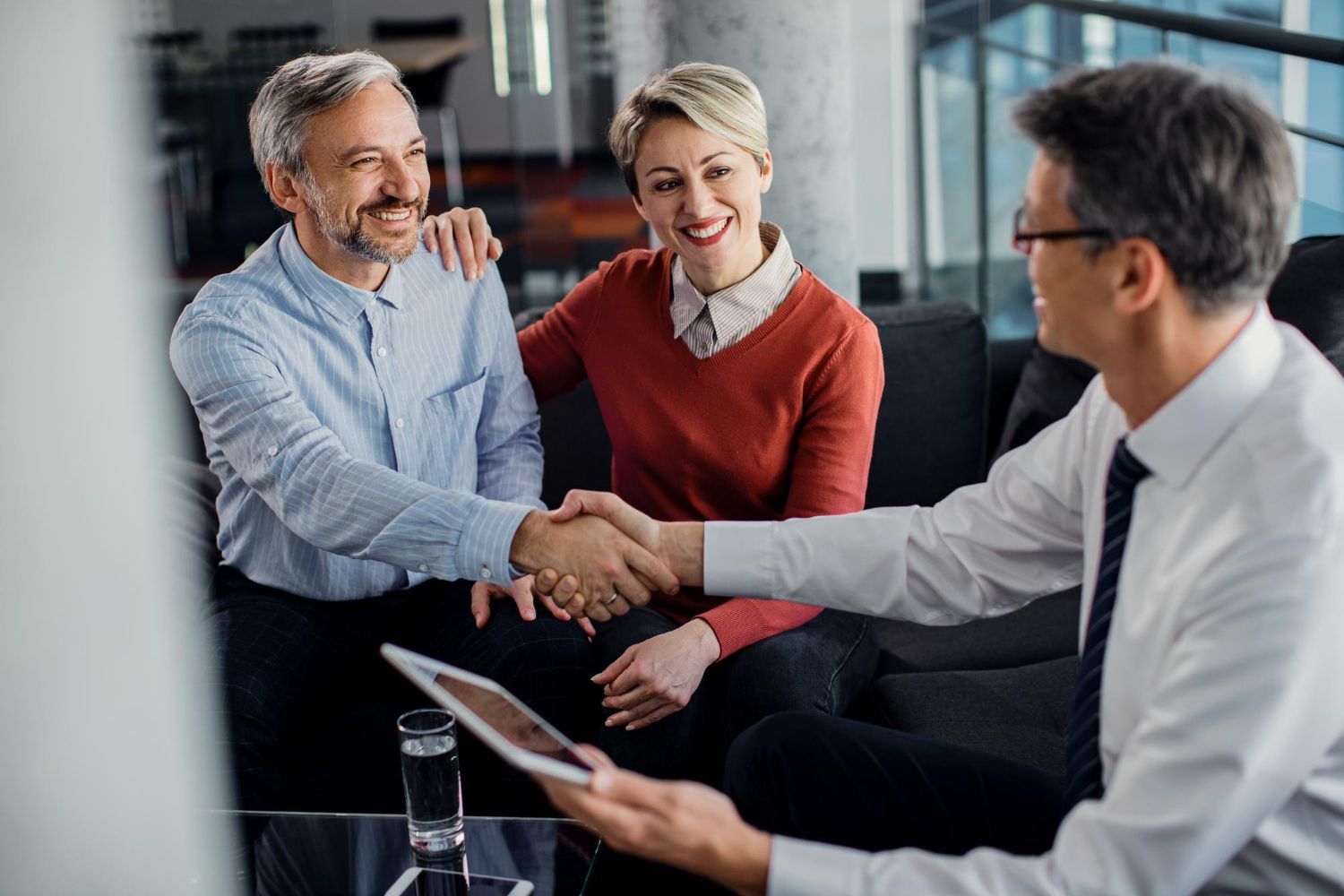 Three coworkers in an office shaking hands during a meeting, with one holding a tablet.