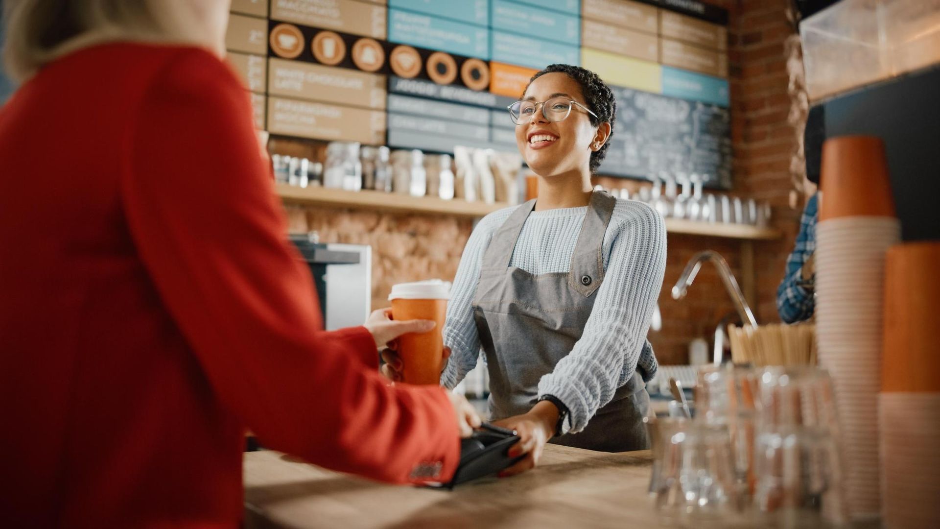 Barista smiling behind a café counter, handing a drink to a customer in a warm, rustic coffee shop