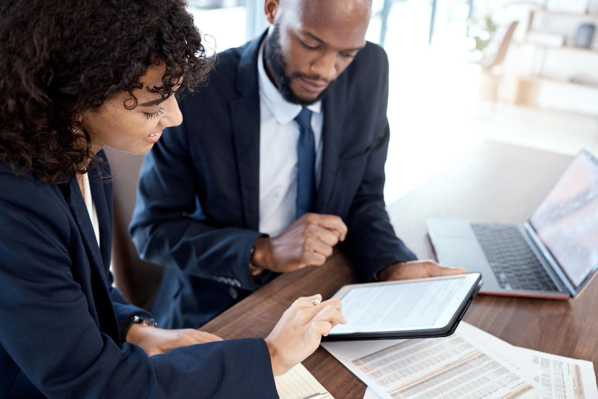 Two coworkers reviewing a tablet and laptop at a desk in a bright office