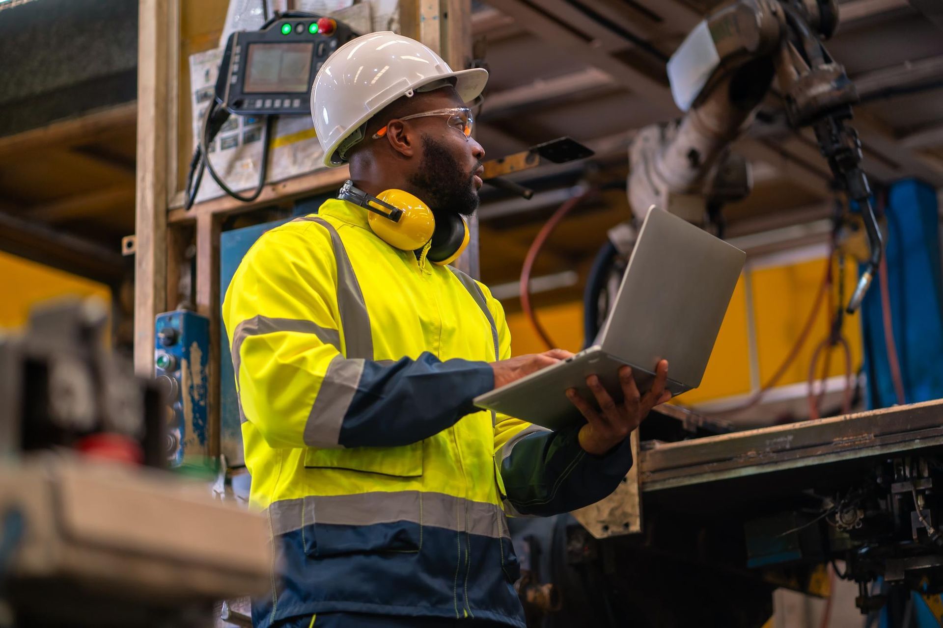 Worker in a hard hat and safety vest uses a laptop in an industrial workshop.