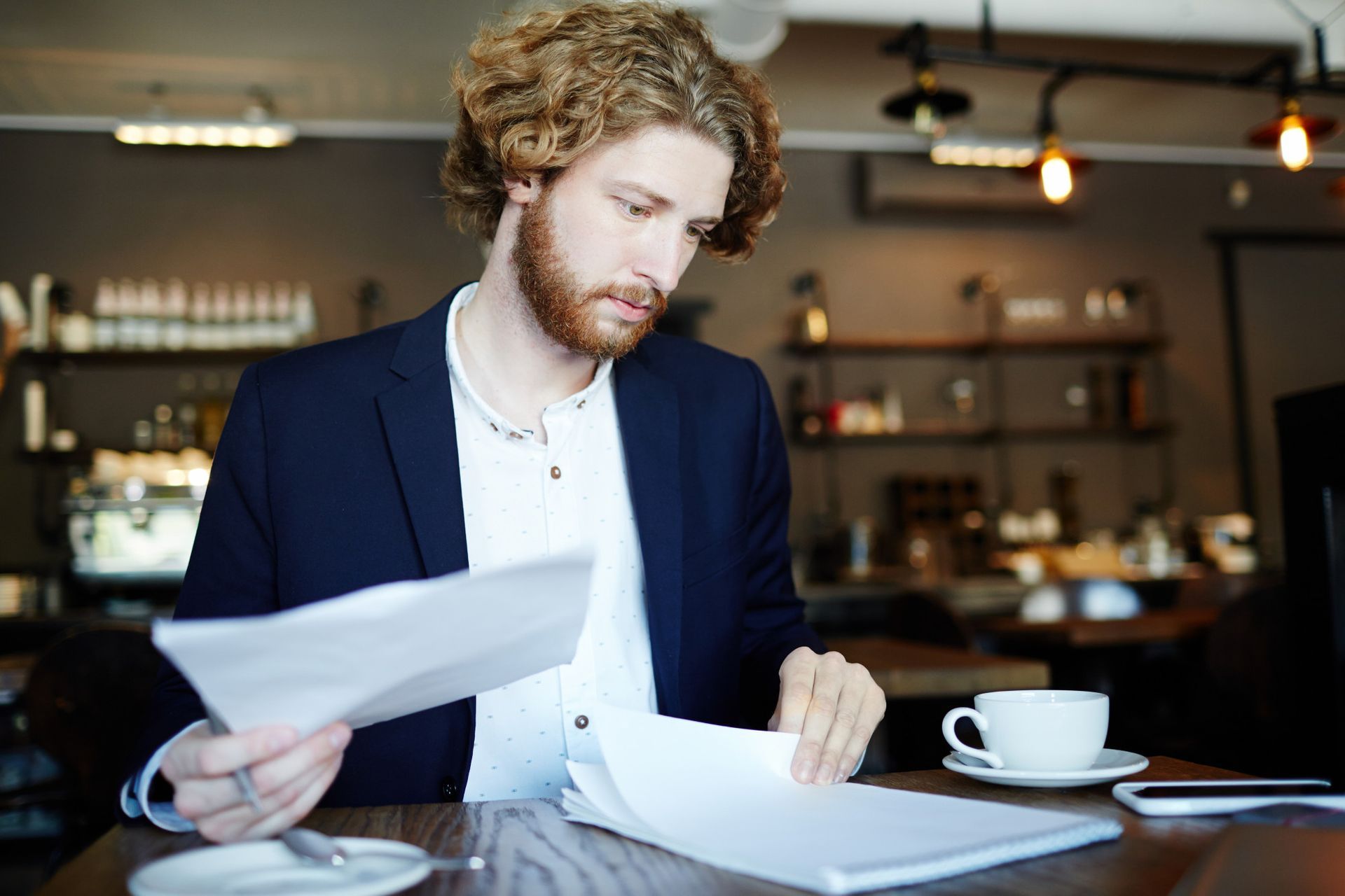 Person reviewing papers at a café table with a cup of coffee and laptop nearby