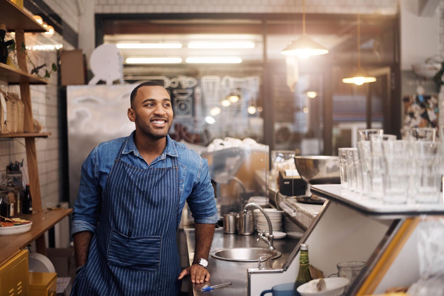 Barista in a blue apron working behind a cafe counter with coffee equipment and glassware