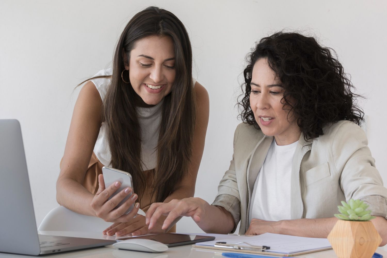 Two women smiling and looking at a phone while working at a desk with a laptop and papers