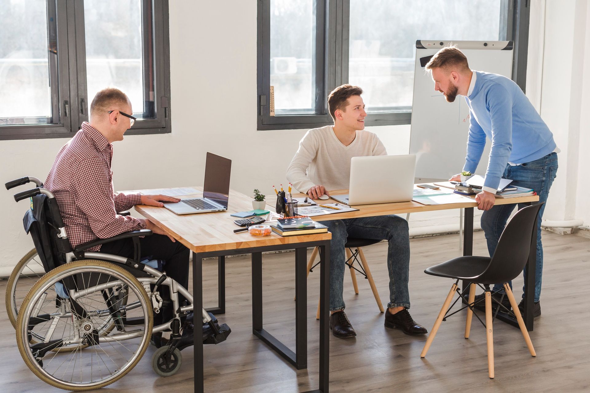 Three coworkers meet in a bright office, one in a wheelchair, discussing work around laptops and a desk.