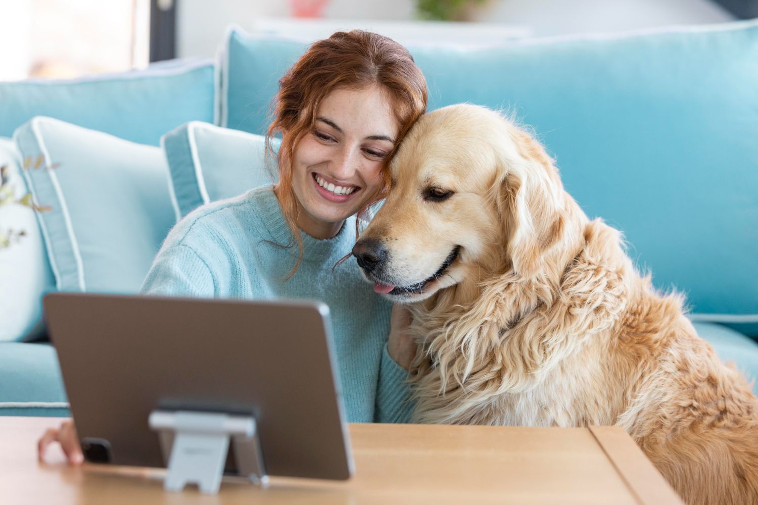 Woman smiling beside a golden retriever on a couch, looking at a laptop on a table