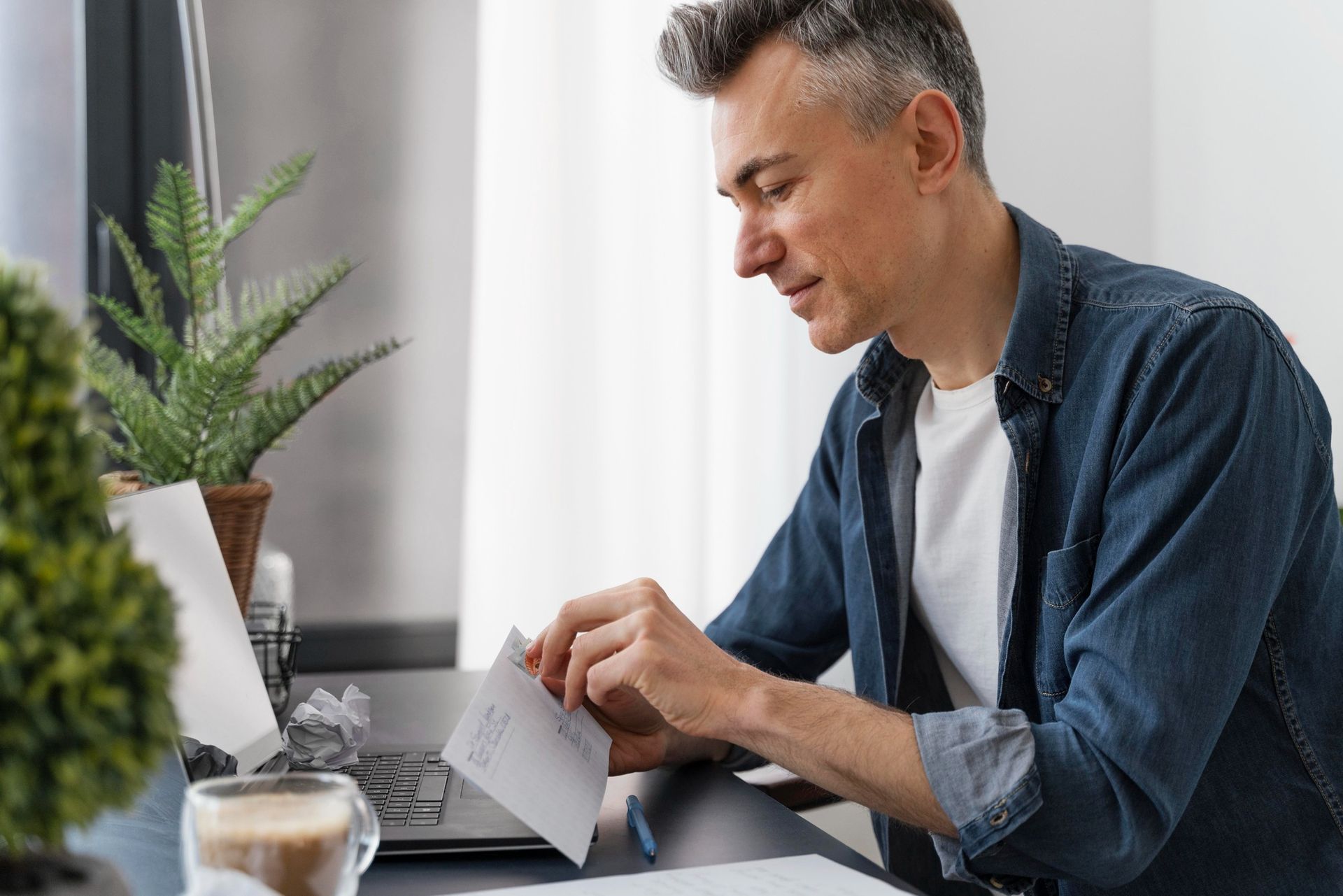 Man working on a laptop at a desk beside a potted plant, in a bright home office