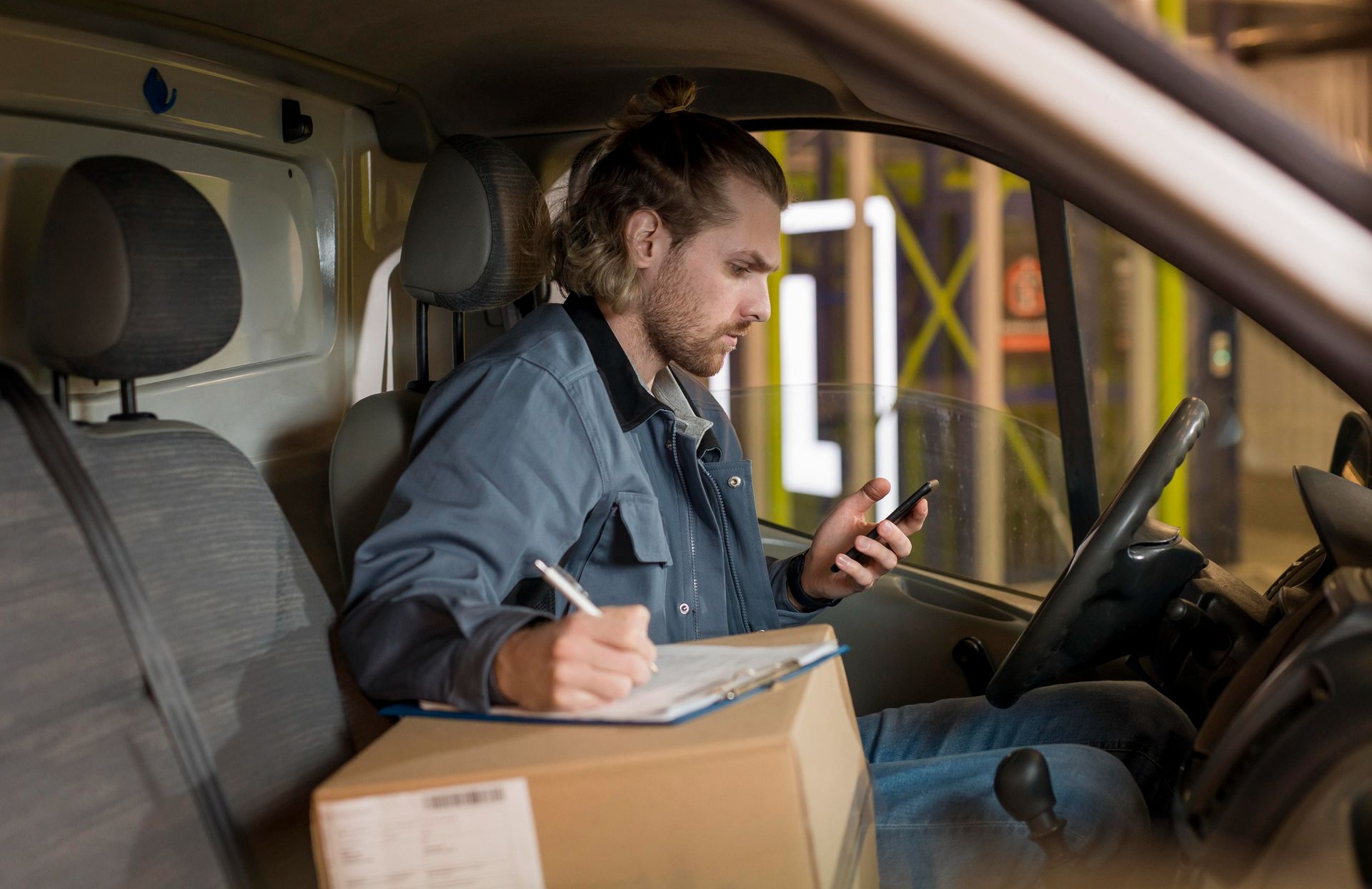 Courier in a delivery van writing on a clipboard while checking a phone, with a package beside him.