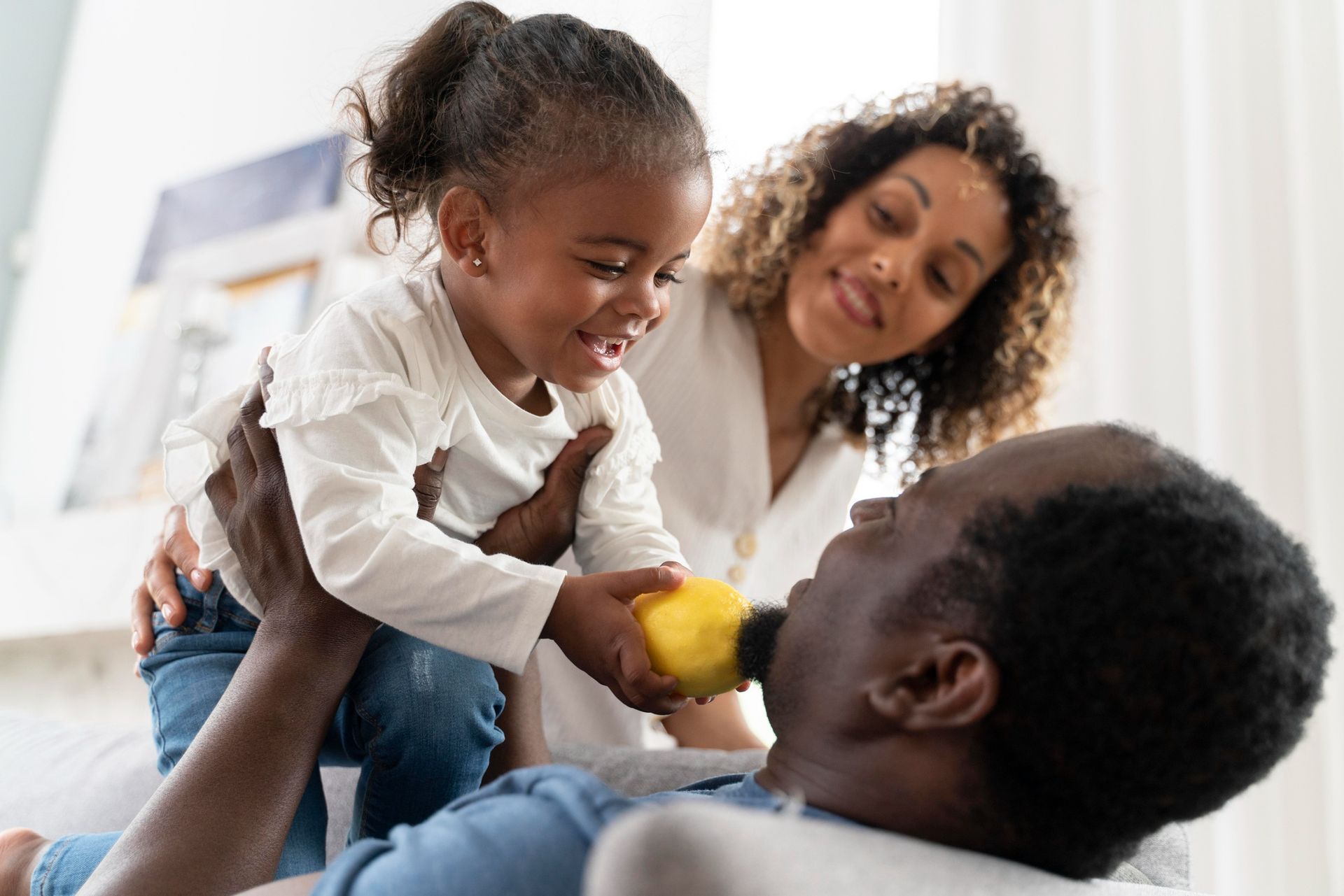 Adult and child smiling on a couch, sharing a yellow fruit in a bright living room