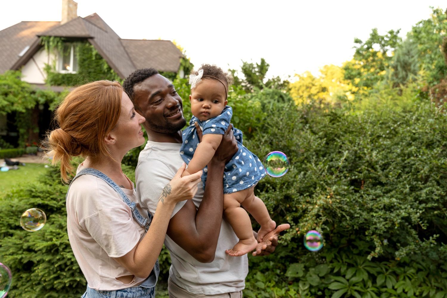 Two adults holding a baby in a garden, with a house and greenery in the background.