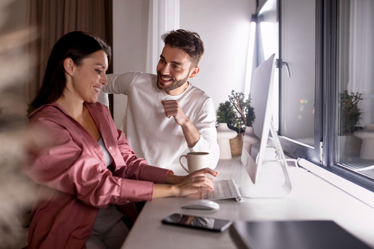 Two coworkers smiling at a desk by a window, sharing coffee and looking at a computer screen