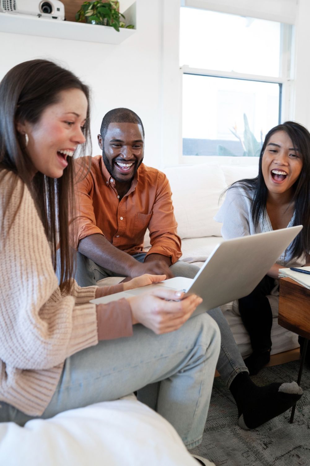 Three friends laughing on a couch while looking at a laptop in a bright living room