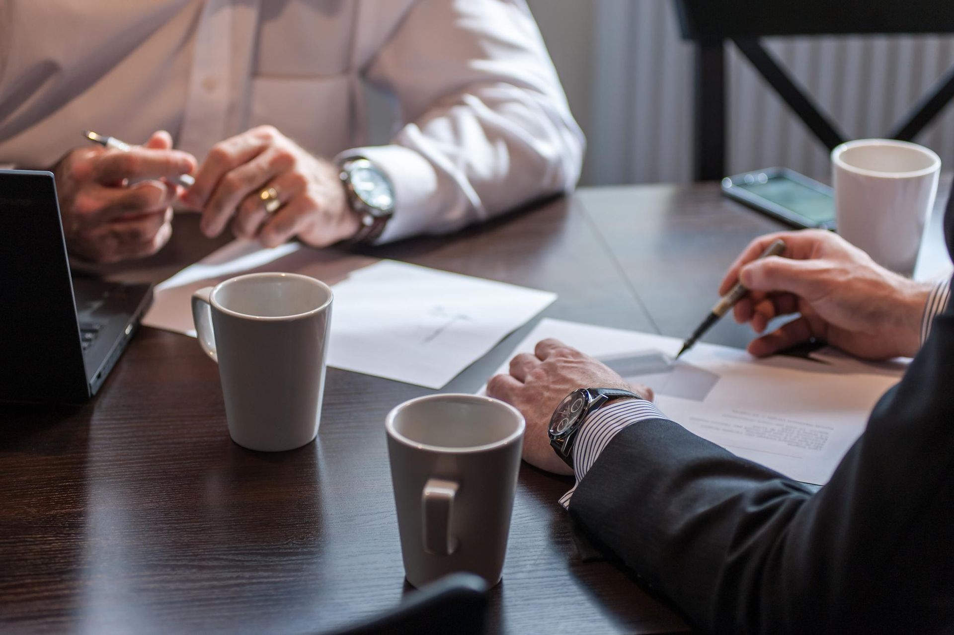 Three people’s hands around a table during a meeting, with papers, coffee cups, and a laptop.