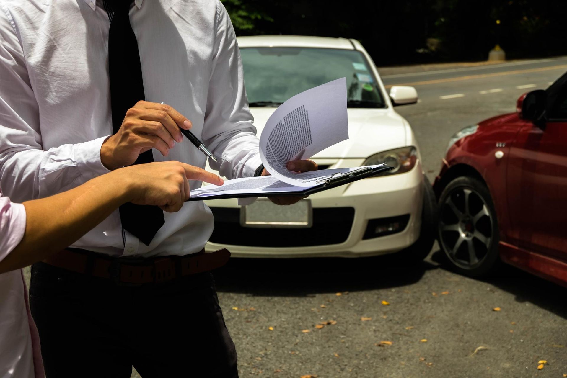 Two people exchanging papers beside parked cars on a sunny street