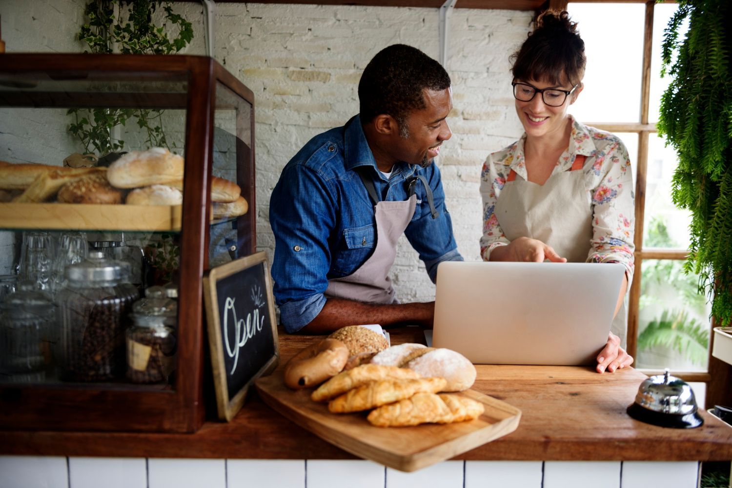 Two people chatting at a bakery counter beside a laptop and fresh bread loaves