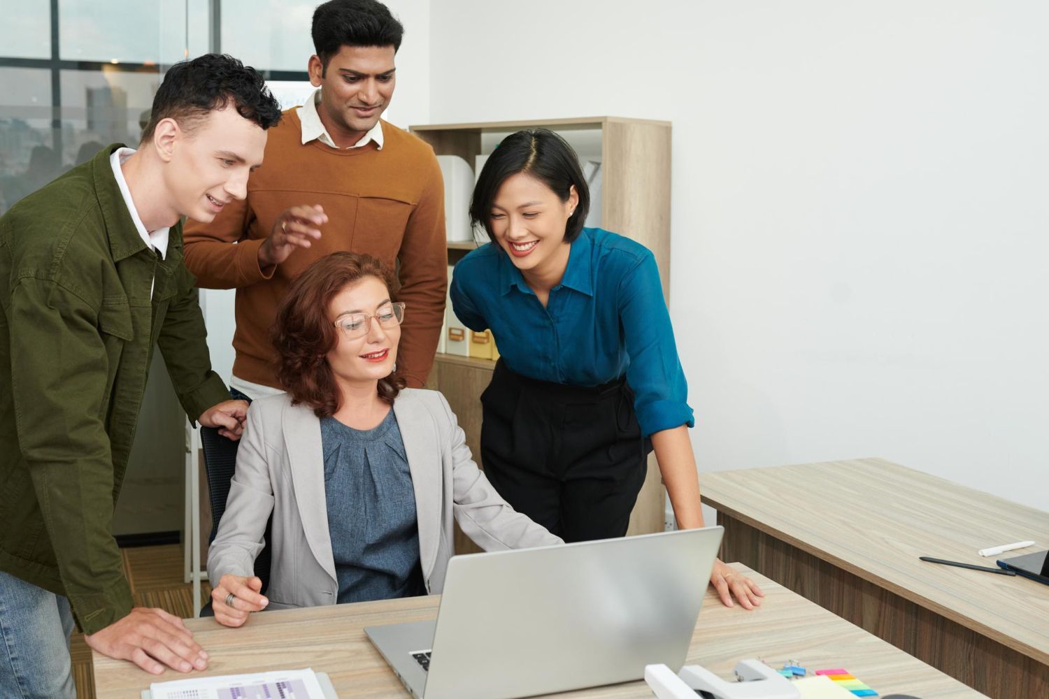 Four coworkers gathered around a laptop in a bright office, smiling and collaborating.