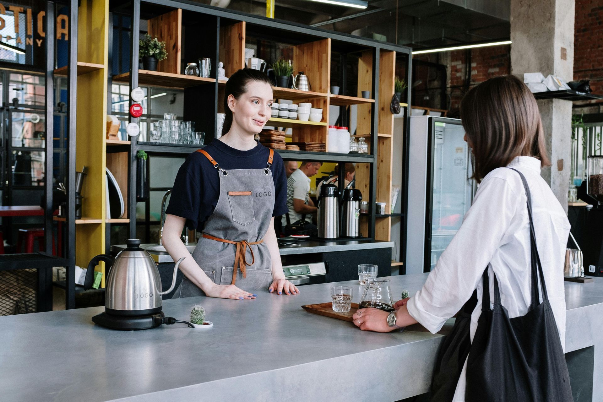 Barista serving a customer at a café counter, with a kettle and pastries in the foreground.