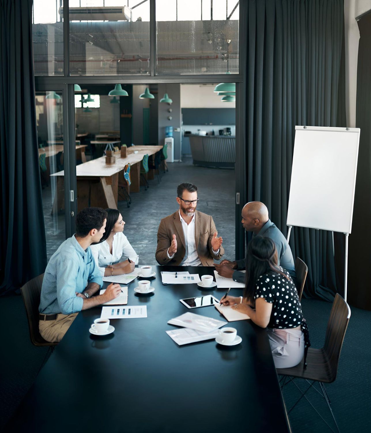 Team meeting around a conference table in a modern office, with papers, laptops, and a blank flip chart.