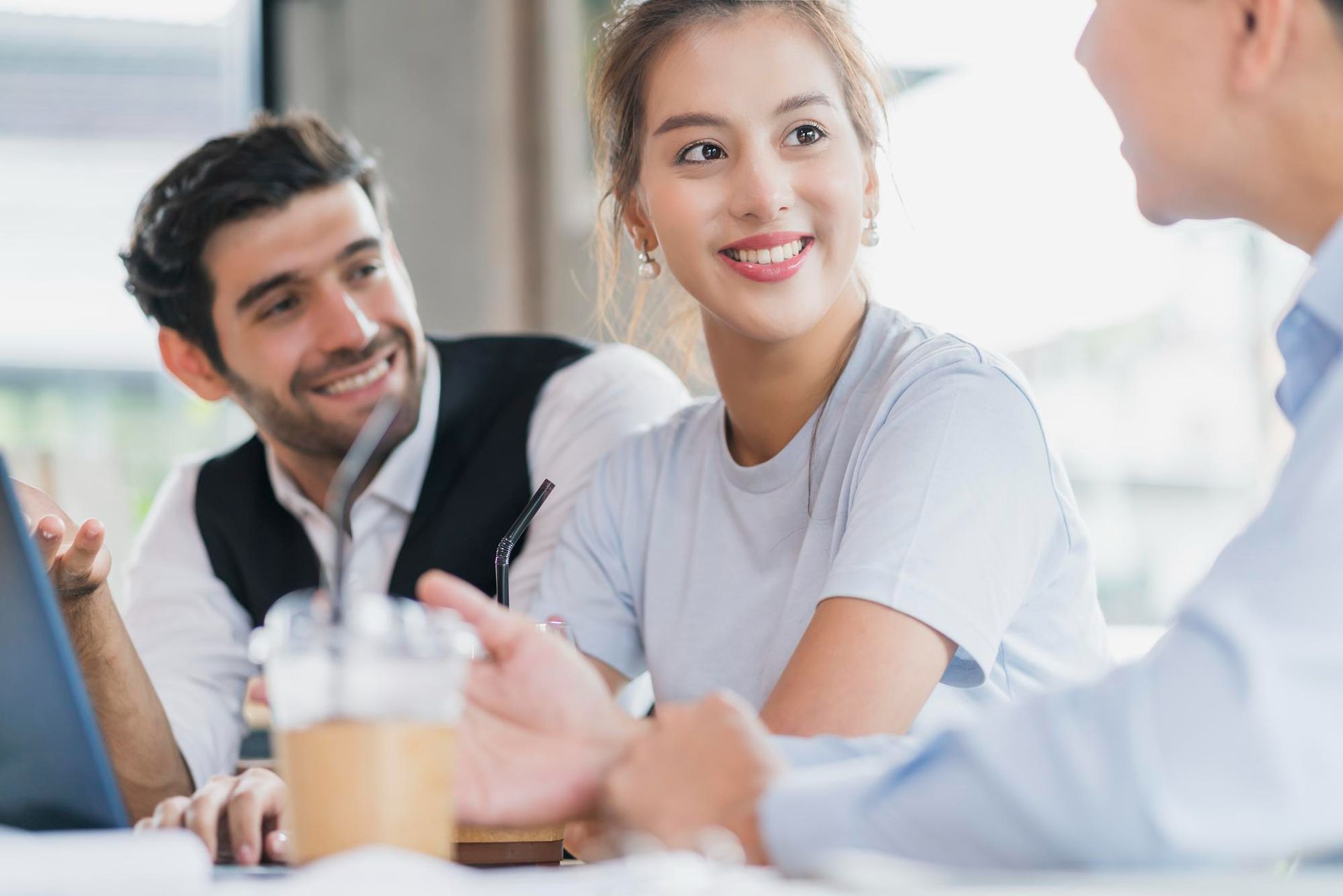 Three coworkers smiling and talking at a table in a bright office meeting.