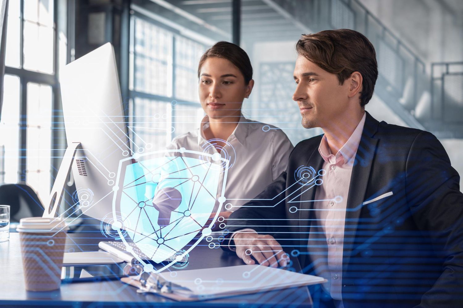 Two coworkers discussing data over a laptop, with a glowing digital shield overlay in a modern office.