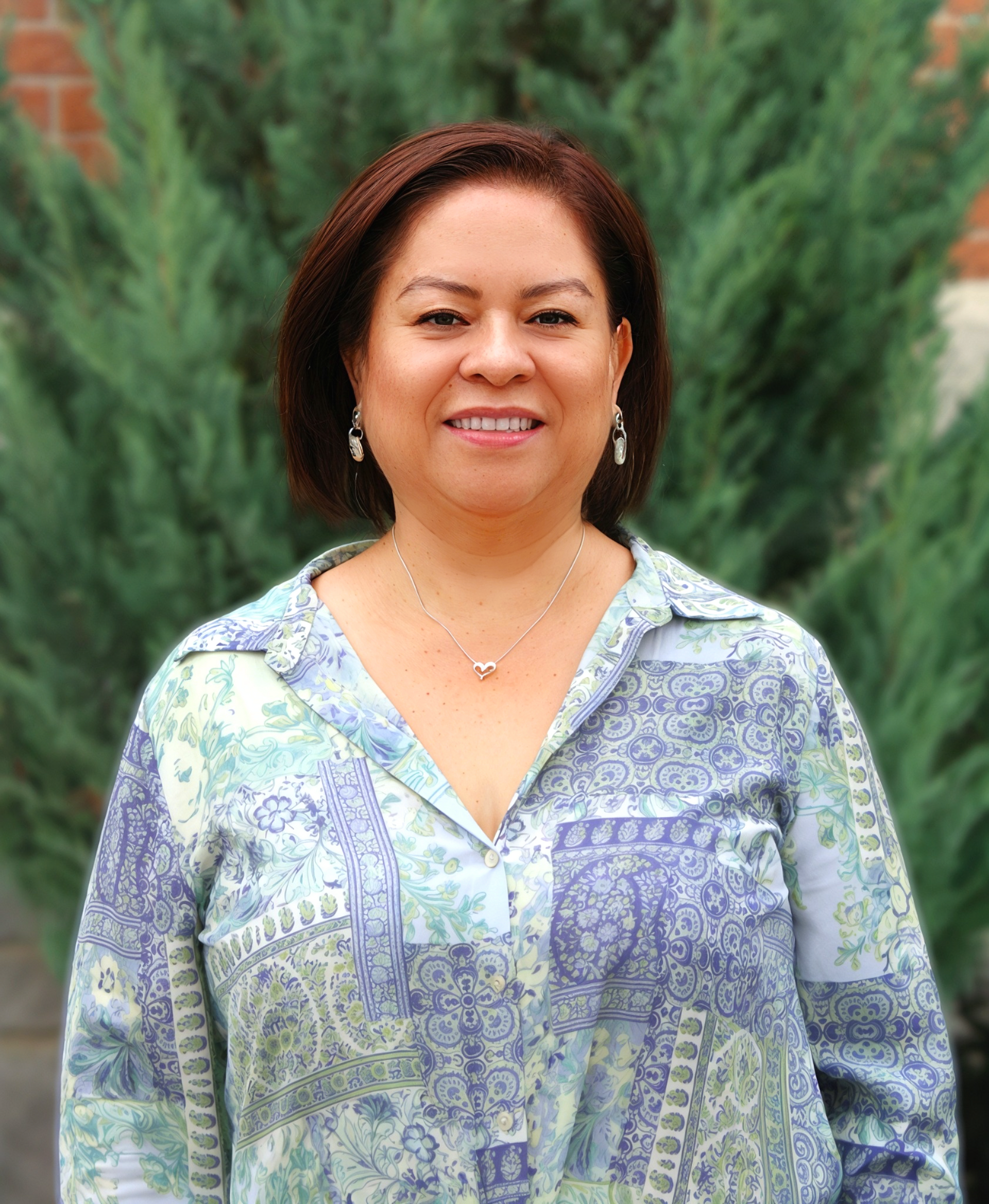 Woman smiling outdoors in a patterned blue-green blouse with trees in the background