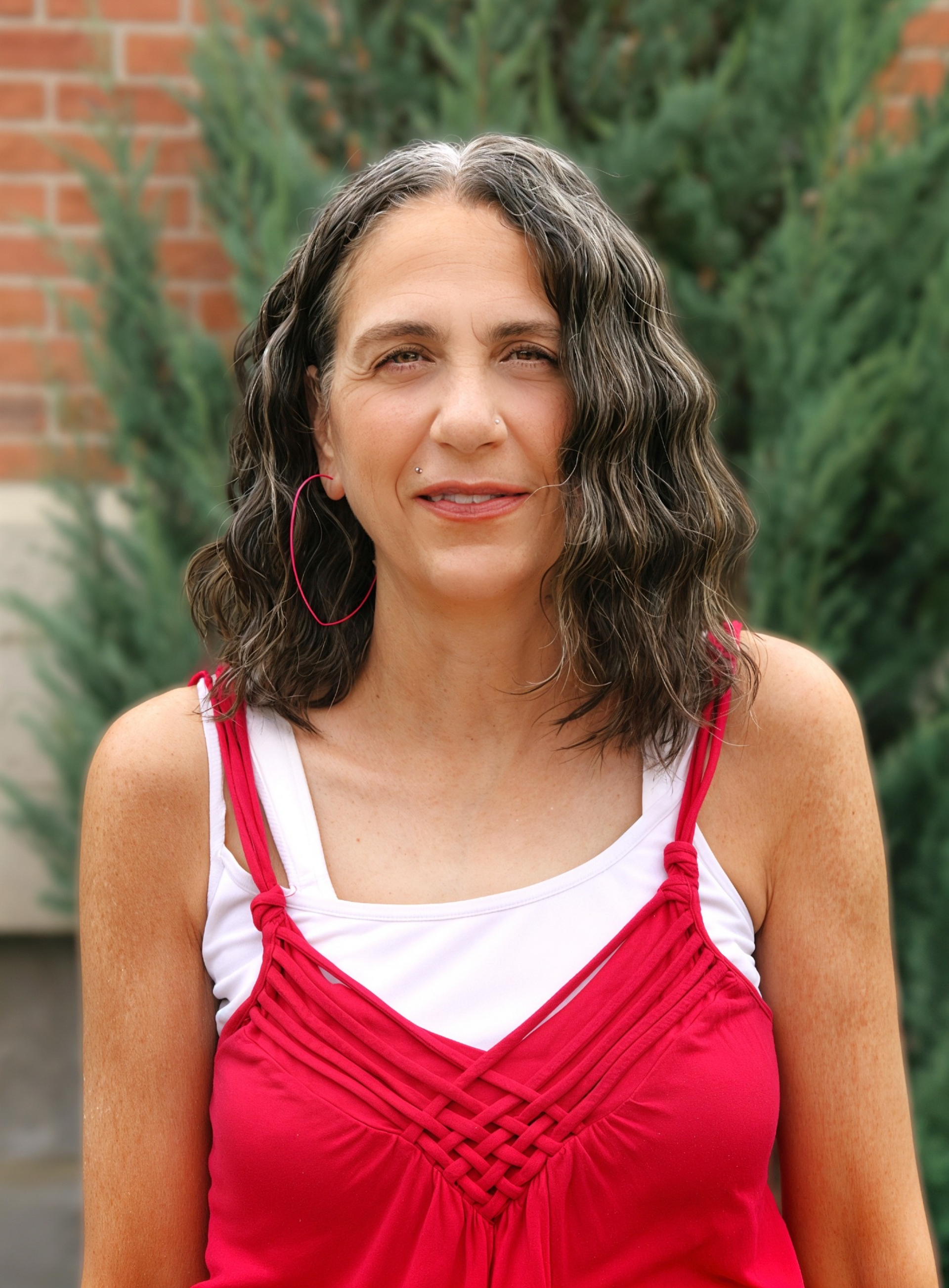 Woman with curly dark hair in a red dress standing outdoors in front of greenery and brick wall