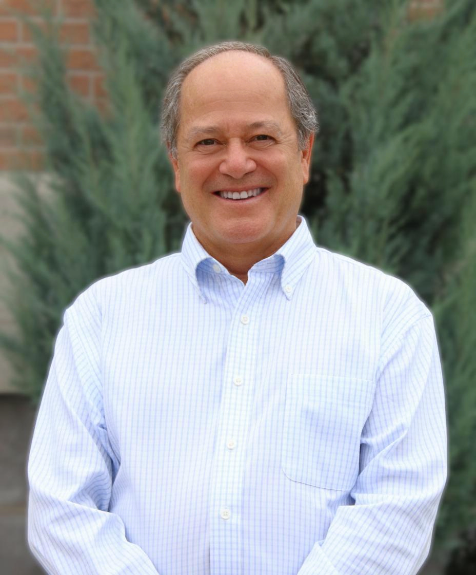Smiling man in a light blue striped shirt standing outdoors in front of greenery