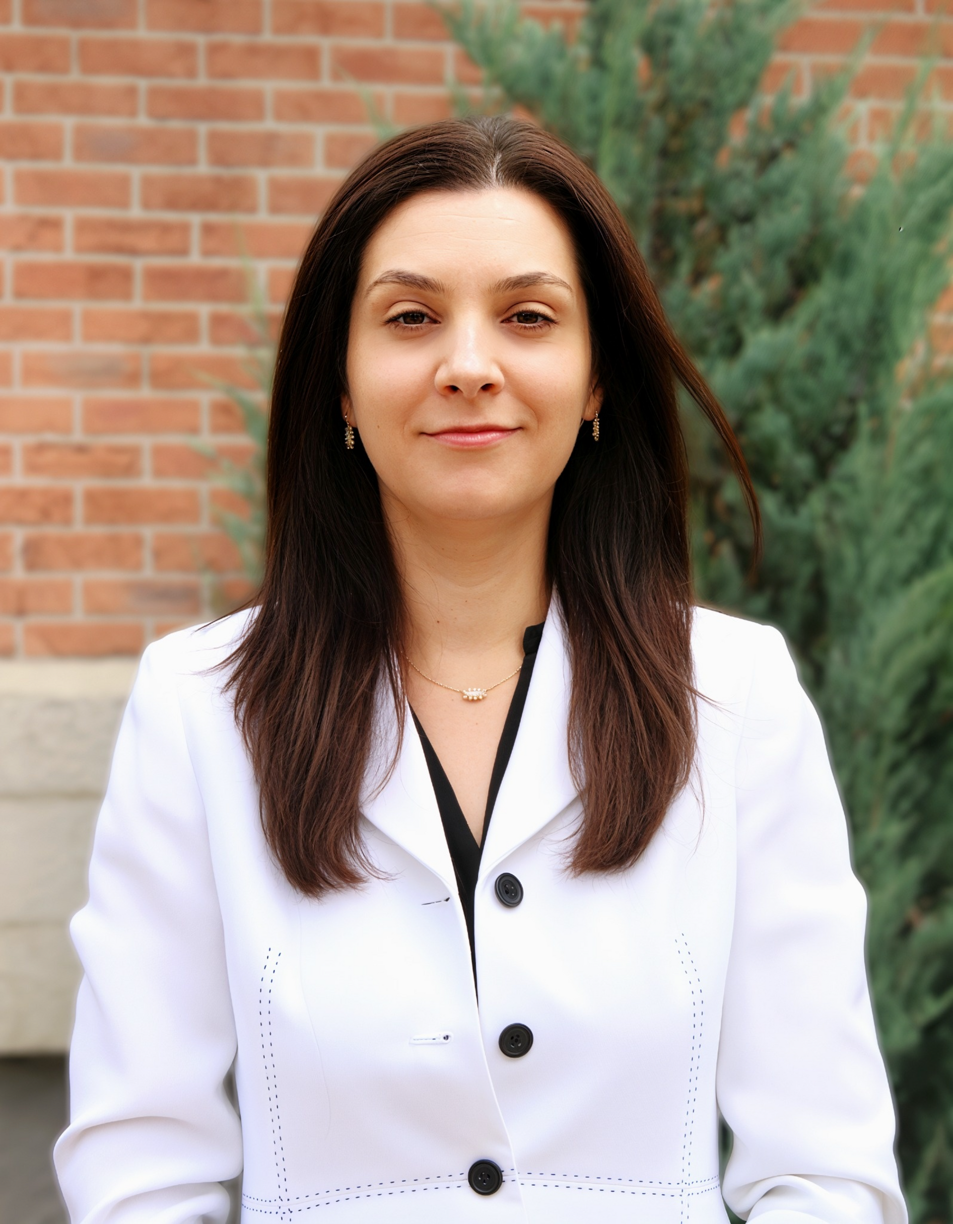Woman standing outdoors in a white blazer, in front of a brick wall and greenery.