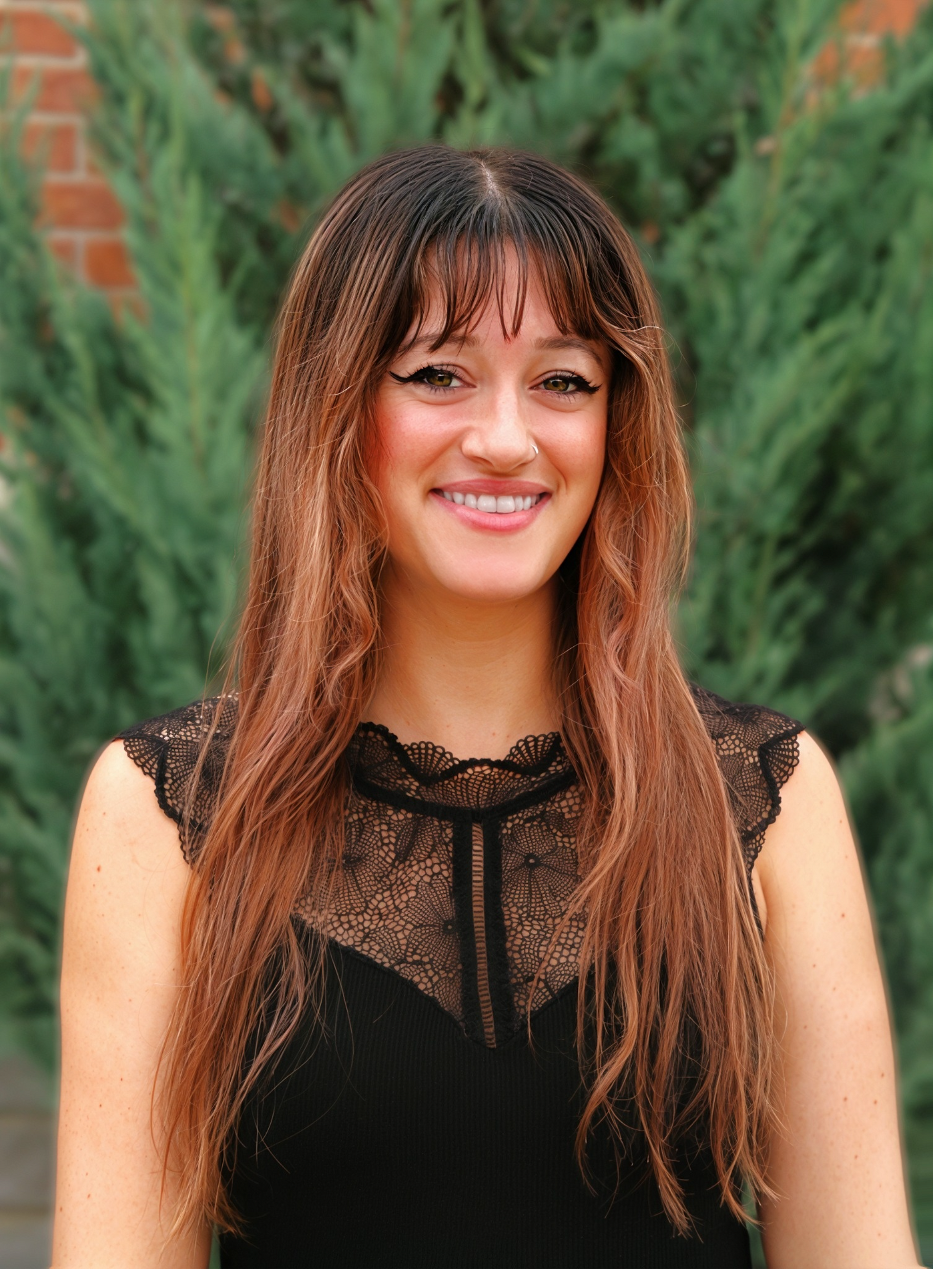 Smiling woman in a black lace top standing outdoors against greenery and brick.