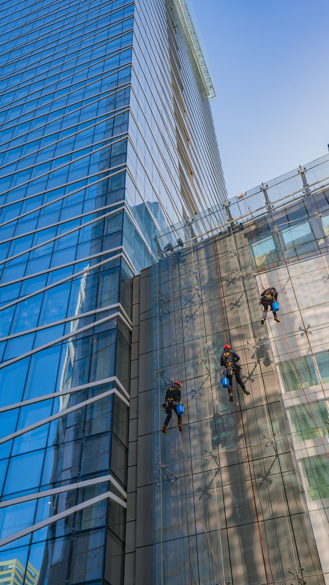 Window washers on ropes beside a glass skyscraper with blue sky reflection