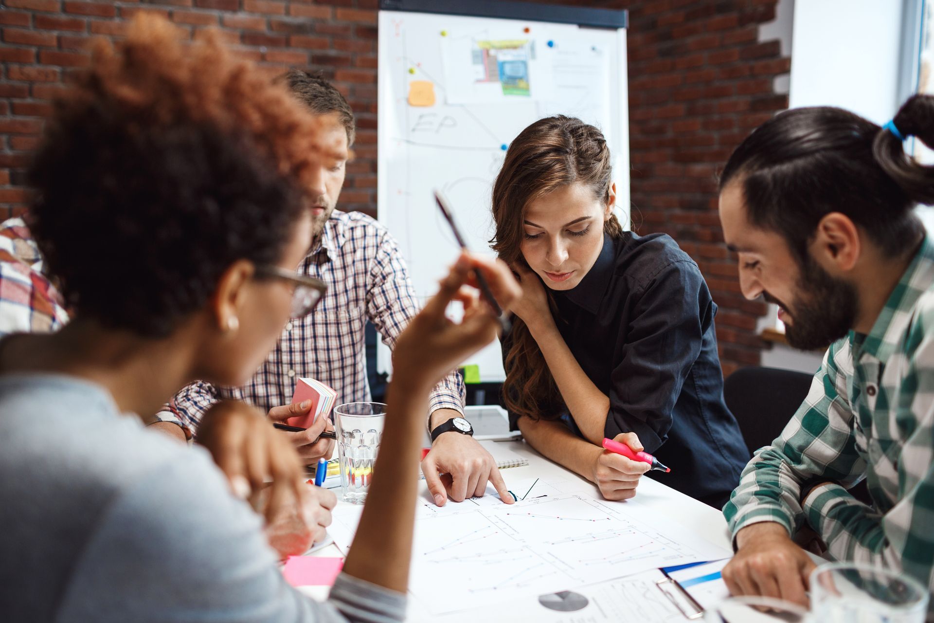 People collaborating around a table, reviewing notes and pointing at a whiteboard in a meeting room.