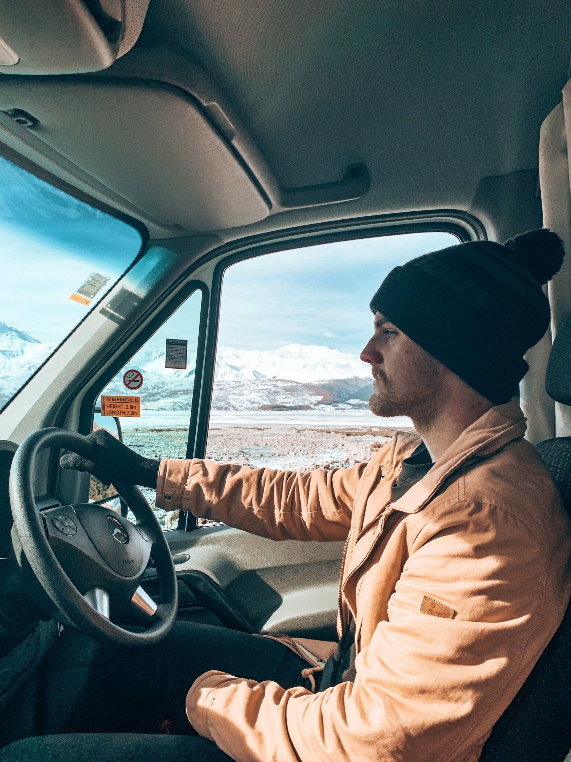 Driver in tan jacket and black beanie inside a van, looking out at a snowy mountain landscape.