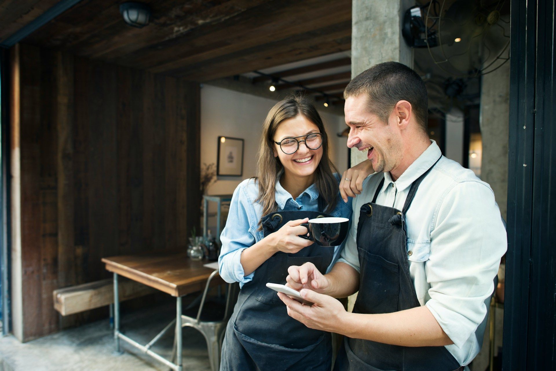 Two café staff in aprons smiling while using a smartphone near the counter