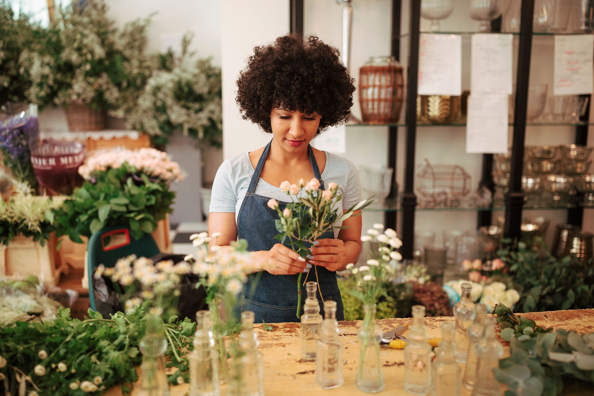Person arranging flowers at a market stall with jars and bouquets in a bright shop setting