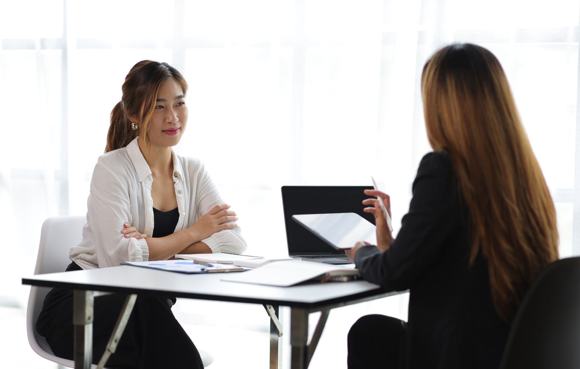 Two women in a bright office meeting at a table with papers and a laptop