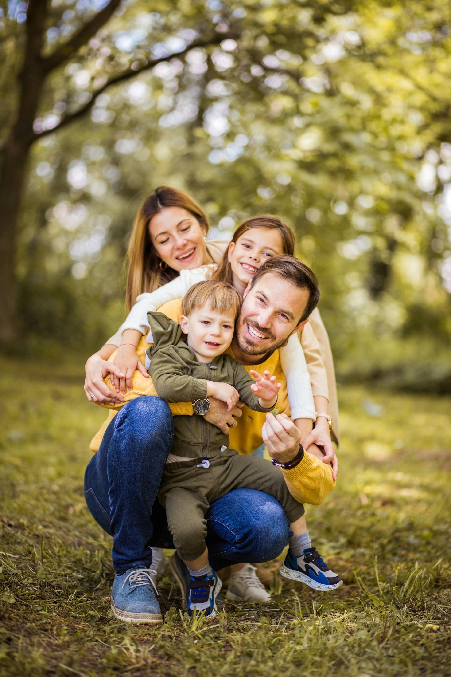 Family of four sitting on grass in a sunlit park, smiling and hugging together