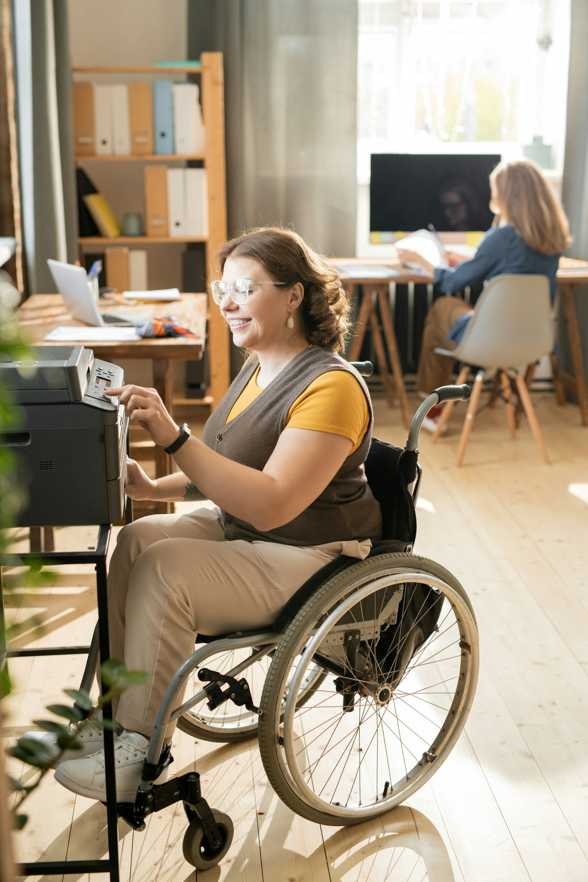 Woman in a wheelchair working at a desk in a bright office, with another person seated in the background.