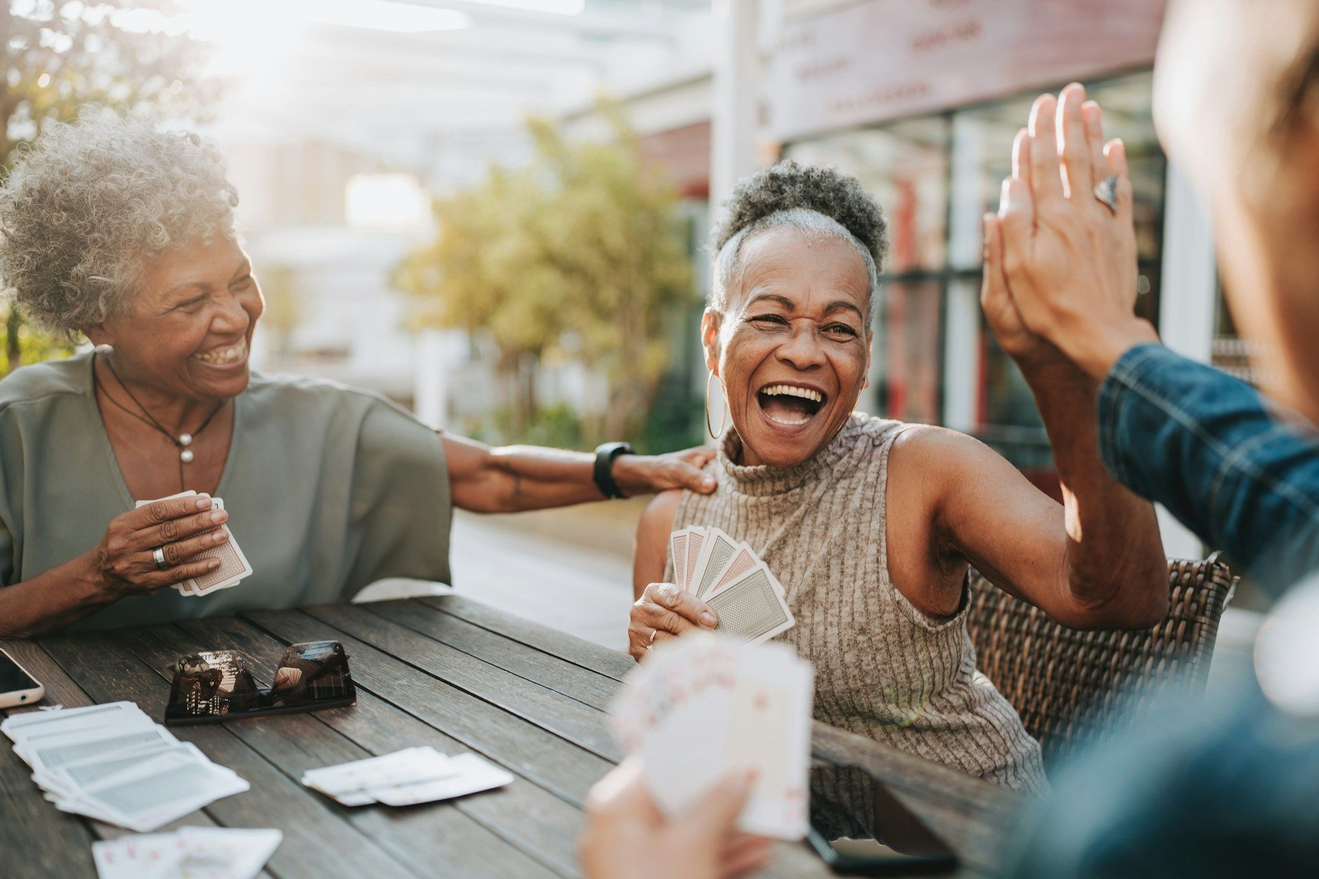 Friends laughing and high-fiving at an outdoor café table with drinks and cards