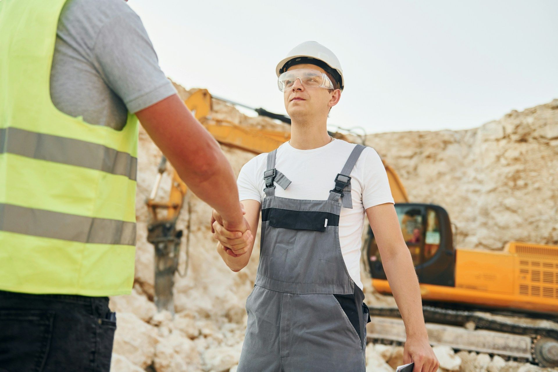 Two construction workers shaking hands at a quarry, with a backhoe in the background