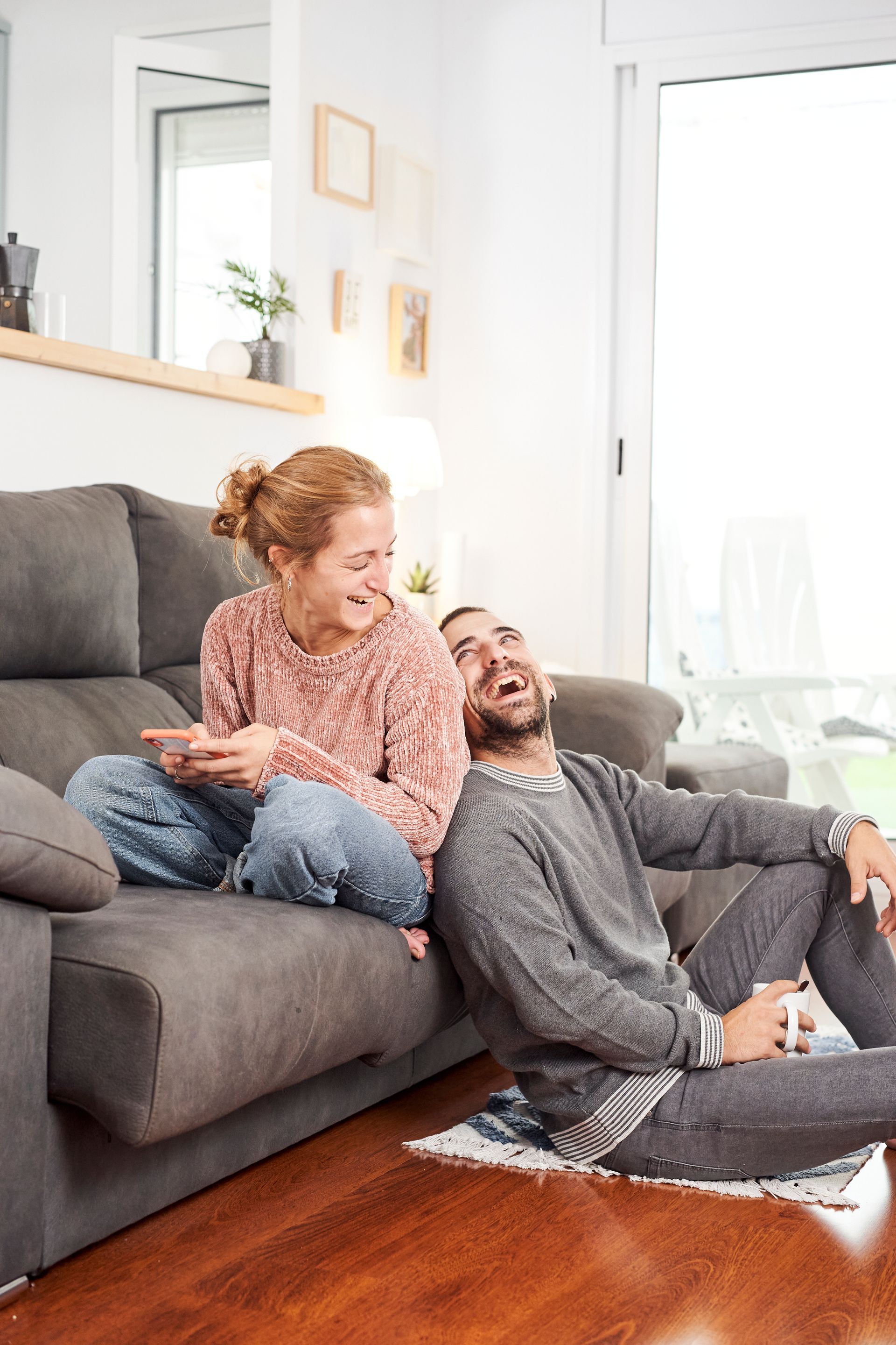 Couple relaxing on a gray couch in a bright living room, smiling and chatting.