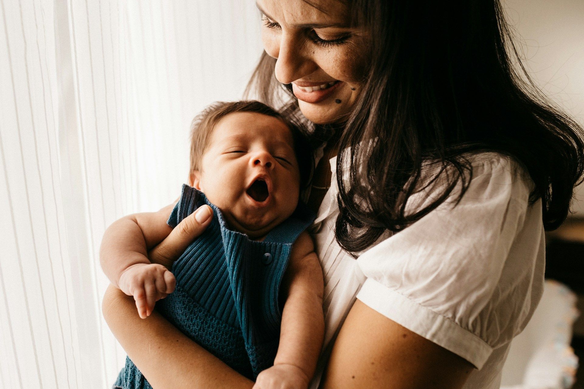 A smiling woman holds a yawning baby wrapped in a blue blanket near a bright window.