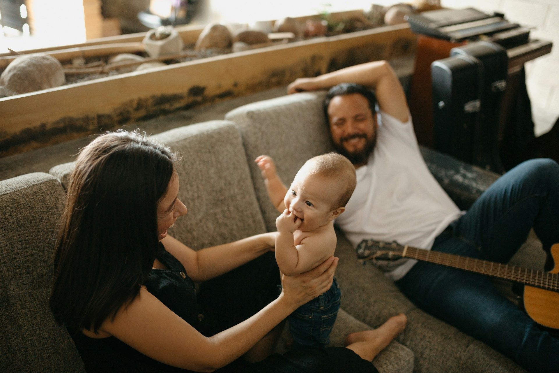 Family sitting on a couch, smiling at a baby, with a guitar nearby in a cozy living room.