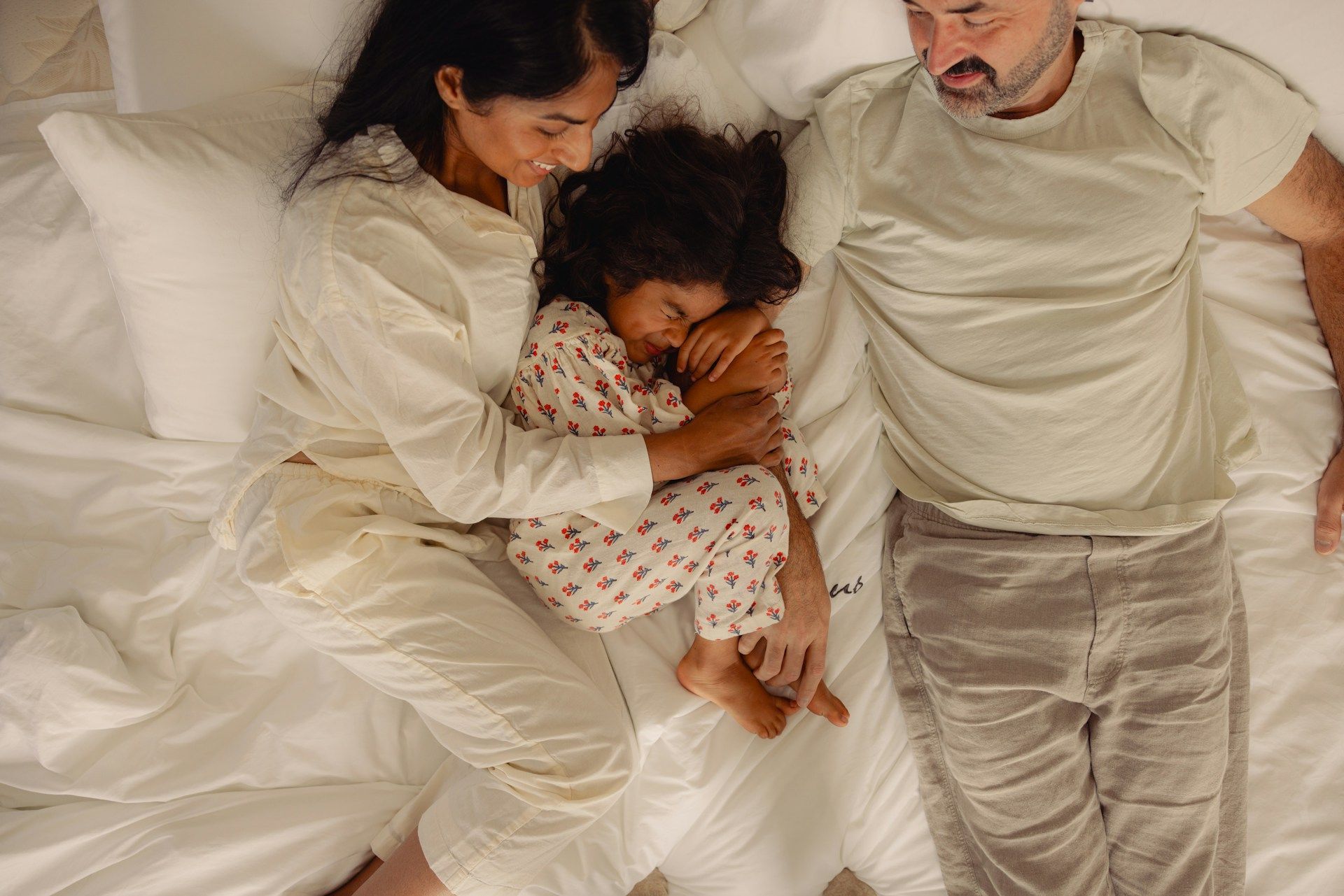 Parents lying on a bed with a sleeping child between them, all in white bedding and pajamas