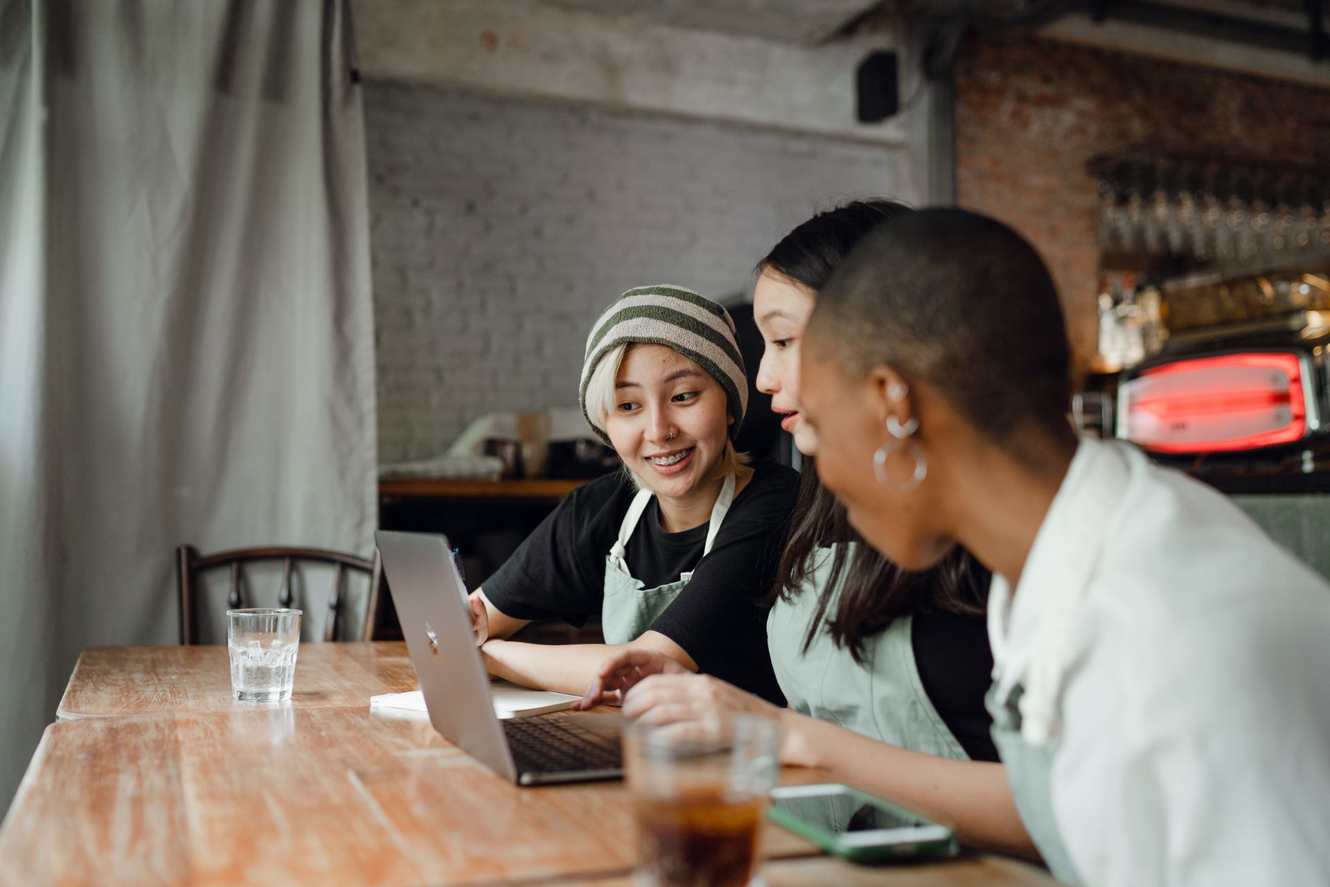 Three coworkers chatting at a laptop in a café, with drinks on the table.