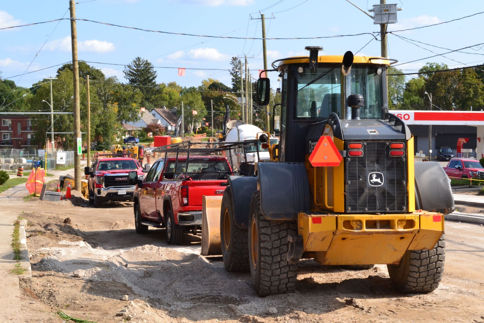 Yellow backhoe and utility trucks on a road construction site, with orange cones and gravel.