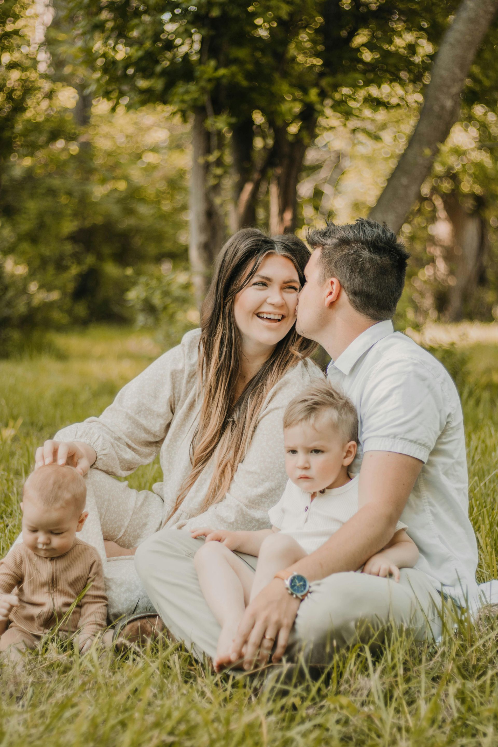 Family sitting in tall grass, smiling together in a sunlit park.