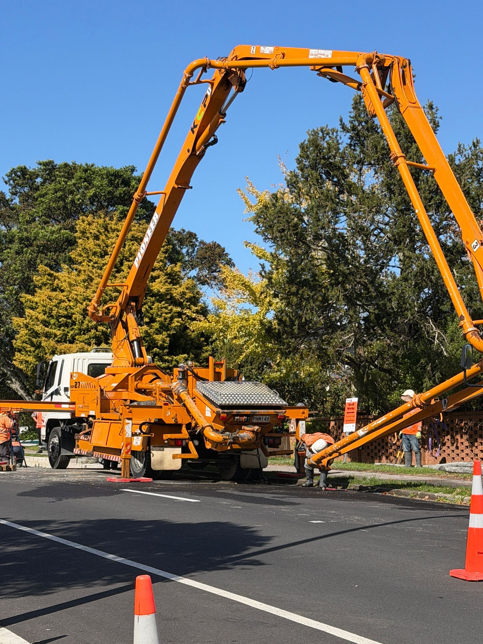 Orange concrete pump truck with long boom extended over a road, surrounded by traffic cones and trees