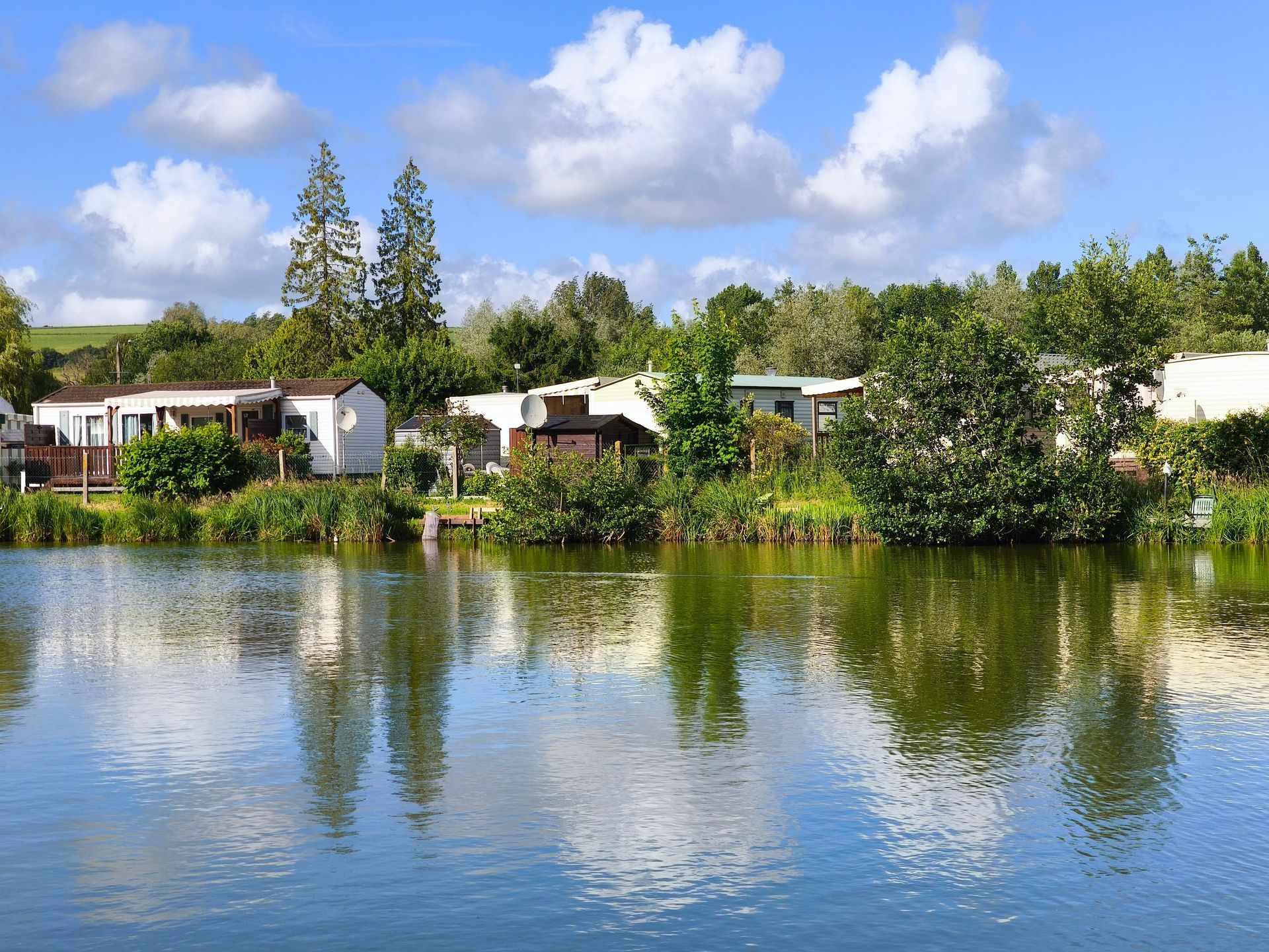 Lakeside homes and trees reflected in calm water under a blue sky with clouds