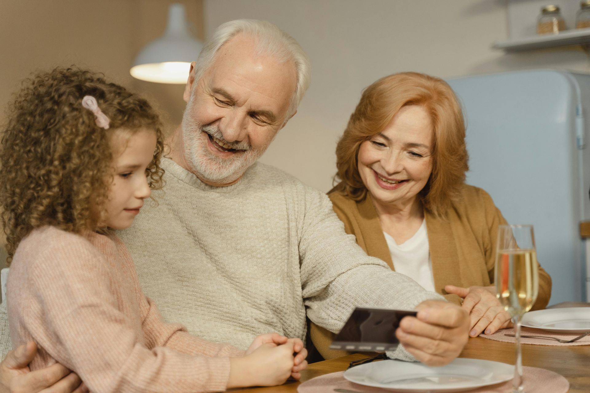 Family sharing a meal at a table, smiling and looking at a phone in a cozy kitchen.